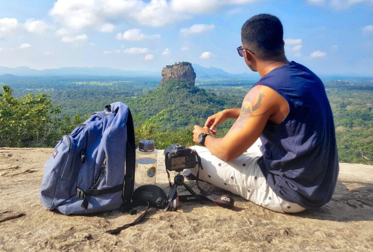 man sitting on top of gray cliff mountain beside backpack water bottle and camera